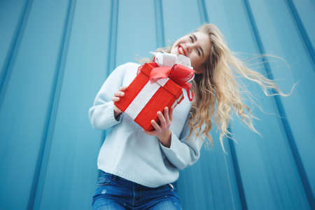 Happy blonde woman in holding gift box and looking at camera while enjoys over blue background. High quality photo.の写真素材