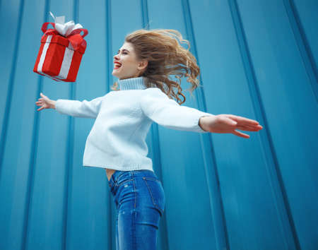 Happy blonde woman in holding gift box and looking at camera while enjoys over blue background. High quality photo.の写真素材