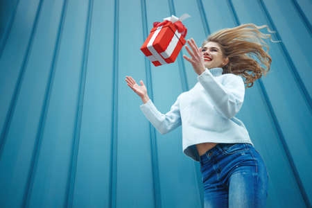 Happy blonde woman in holding gift box and looking at camera while enjoys over blue background. High quality photo.の写真素材