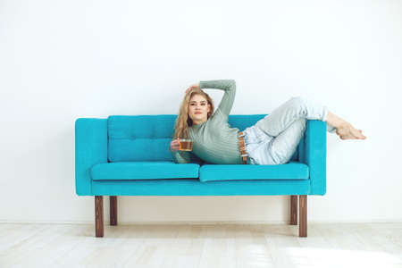 Beautiful woman is drinking tea on the sofa at home. Sportive young girl doing yoga pose on the couch. High quality photo.の写真素材
