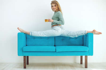 Beautiful woman is drinking tea on the sofa at home. Sportive young girl doing yoga pose on the couch. High quality photo.の写真素材