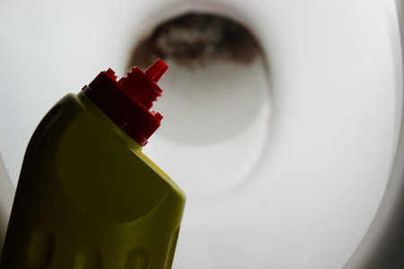 Close-up of a yellow cleaner bottle with a red angled spout aimed at a toilet bowl showing brown...