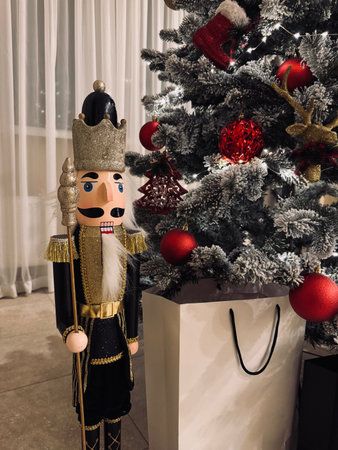 Wooden nutcracker figure standing next to a Christmas tree decorated with red baubles and lights, with a large white gift bag in the foreground. Festive home interior holiday decoration concept.の写真素材