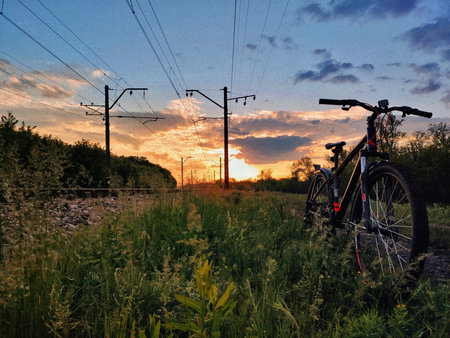 A mountain bike rests beside railway tracks at golden hour, with glowing clouds and tall grass framing an adventurous countryside evening.の写真素材