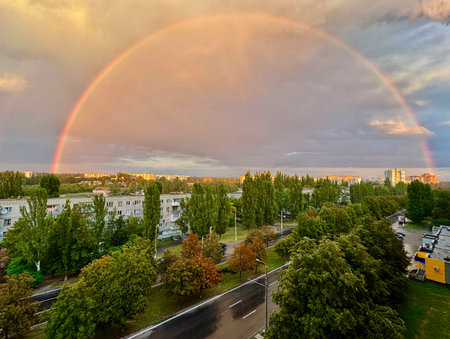 Wide view of complete rainbow spanning across apartment blocks, trees and wet streets after a shower. Soft evening light over urban neighborhood.の写真素材