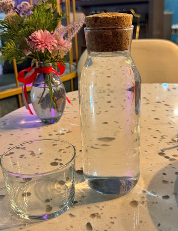 Clear water carafe with cork stopper and matching low glass on a terrazzo table, soft reflections...