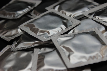Many sealed silver condom packets scattered across a dark surface. Studio still life for contraception, protection, and public health topics.の写真素材