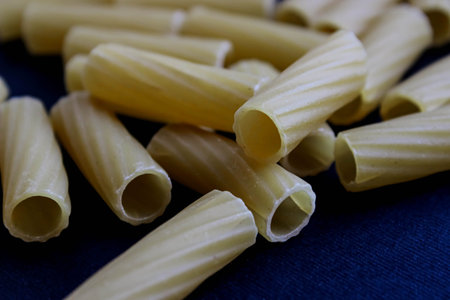 Tight macro of raw rigatoni tubes showing flour dust and ridged texture on a dark surface. Ideal for...