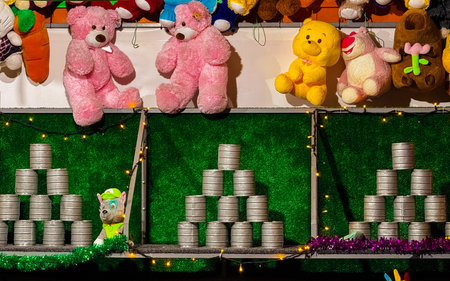 A brightly lit carnival game stand displays stacked metal cans ready for a throwing challenge. Above...