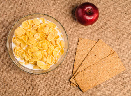 Bowl of cornflakes with milk, crispbreads and apple on sacking backgroundの写真素材