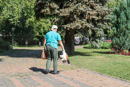 A woman works with a blower to remove the cut grass and leaves from the alley path in the city parkの写真素材