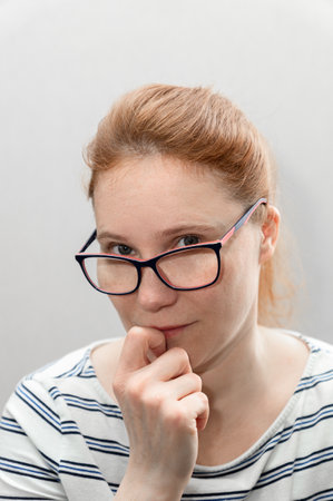 Portrait of a middle-aged woman 48 years old with red hair and freckled on a light blurred background wearing glasses as a mother, teacher, wife, a woman with a hand near her face.の写真素材