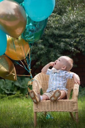 Little boy blond European German age one 1 year old sits in a rattan chair with colored balloons for his birthday.の写真素材