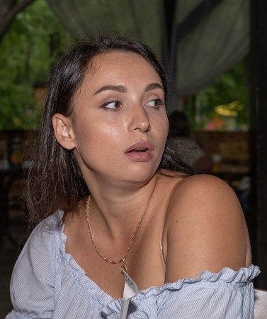 A young pretty woman, age 30, thirty years old, swarthy brunette, looks away in surprise in a summer outdoor restaurantの写真素材