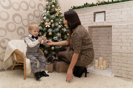 A little boy with his mom sit near the Christmas tree with festive gifts and toys garlands and waits for the coming of Christmas and a magic gift from Santa Claus.の写真素材