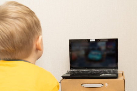 Little boy 3 years old looks cartoon on a black laptop screen via the Internet, close up.