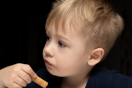 Little boy 3 years old blond white eating a waffle tube on a black background and looking to the...