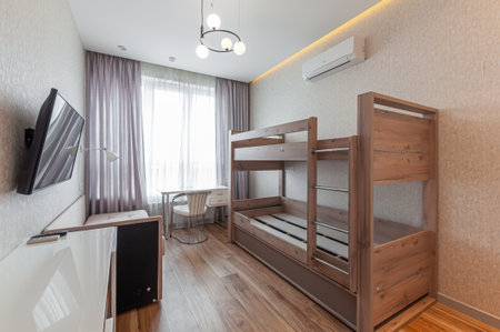 Modern room with a wooden bunk bed, a desk, and a wall-mounted TV. Light interior with wooden flooring and soft lighting creates a cozy atmosphereの写真素材