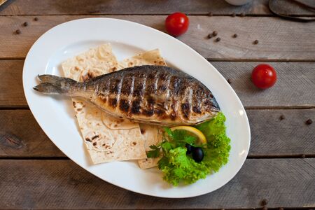 dorado food photo grilled fish trout with vegetables on a white plate with onion halves tomatoes green branches on a blue wooden backgroundの写真素材