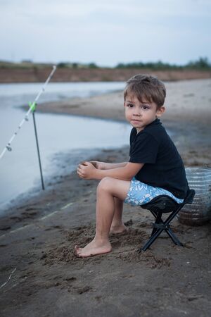 a boy on the Bank of the river is sitting on a chair and fishing. Family vacations, hobbiesの写真素材