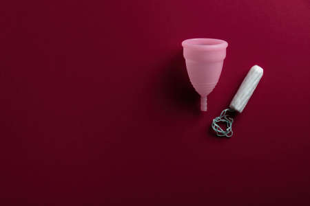 An eco-friendly, reusable pink menstrual cup made from soft medical grade silicone sits next to a new cotton disposable tampon. On a dark red blood-colored backgroundの写真素材