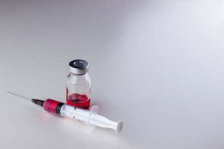 Red liquid (vitamin B12) in a disposable syringe with a needle and in a glass ampoule on a white...