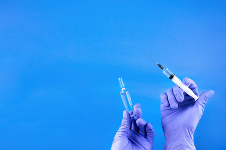 a disposable syringe with a transparent needle and an ampoule of liquid in male hands in blue (lilac) nitrile gloves. On a blue background, lots of empty spaceの写真素材