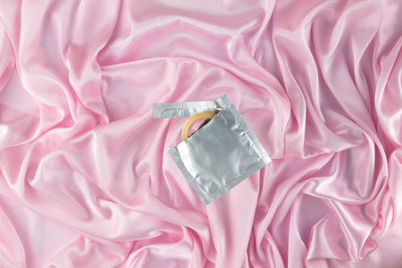 A piece of a condom sticks out of a foil package against a pink silk sheet. Safe sex and reproductive health concept.の写真素材