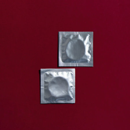 Two condoms in foil packaging lie on a dark red background. Safe sex and reproductive health concept.の写真素材