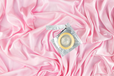 An unpacked condom lies on a foil package against the background of a delicate pink silk sheet. Safe sex and reproductive health concept.の写真素材