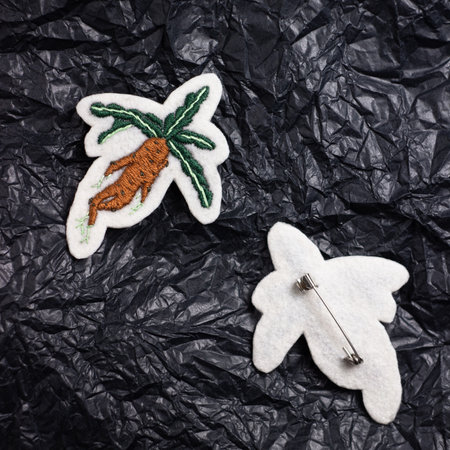 Brooch embroidered with threads on white felt in the form of a mandrake root, next to it lies its back side with a pin. Against the background of crumpled black paperの写真素材