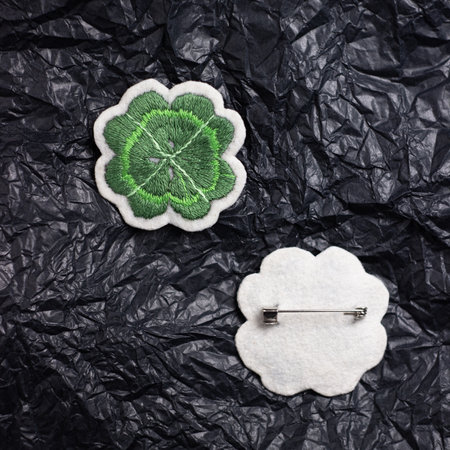 Brooch embroidered with threads on white felt in the form of a clover, next to it lies its back side with a pin. Against the background of crumpled black paperの写真素材