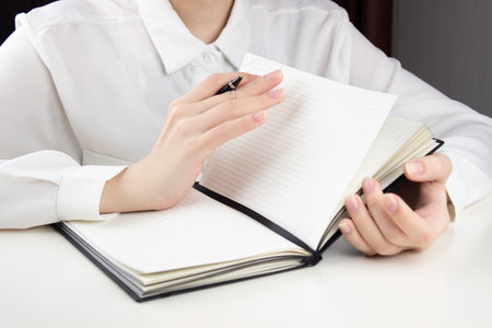 A young girl flips through the pages in a notebook, a pen in her hand. At the white tableの写真素材