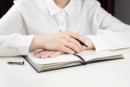 A young girl sits at a table with her hands folded on an open notebook, a pen in her hand, on a white tableの写真素材