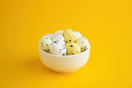 Several quail eggs lie in a white ceramic bowl on a yellow background. Lots of empty space, Easter...