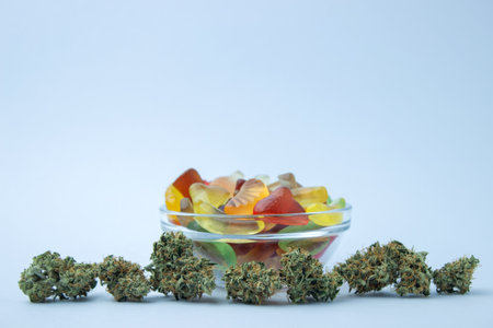 Dried cannabis buds lie in a line in the foreground, behind a small glass bowl of gummy candies. On a pale blue background, lots of empty spaceの写真素材