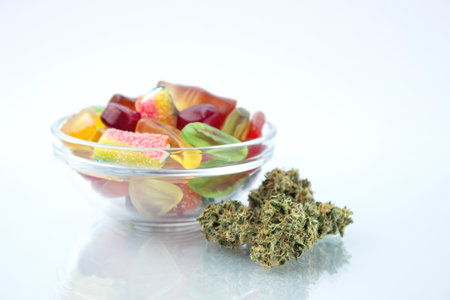 Assorted different gummy candies in a small glass bowl, near in the foreground several buds of dried medical marijuana, reflected on the glassの写真素材