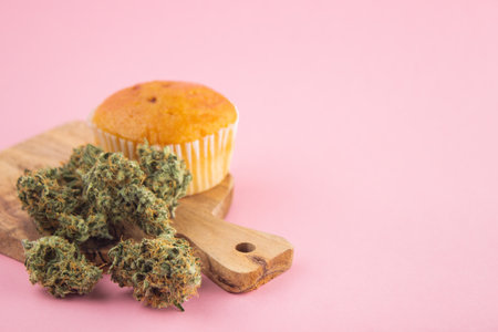 On a wooden board, a cupcake with the addition of cannabis, dry marijuana buds in the foreground. On a pink background, a lot of empty spaceの写真素材