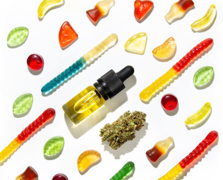 Among Chewing marmalade of different shapes, tastes and colors, a dried cannabis bud and bottles of marijuana seed oil lie in a pattern on a white background in the center.の写真素材
