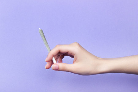 A woman's hand with natural nails holds a joint with medical marijuana on a light purple background. copy spaceの写真素材