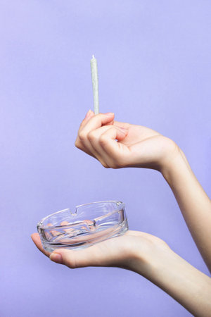 Female hands with natural nails hold a joint with medical marijuana and an empty glass ashtray on a light purple background. copy spaceの写真素材