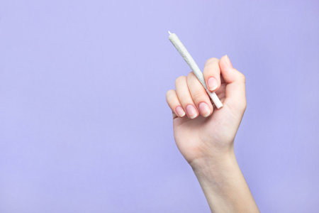 A woman's hand with natural nails holds a joint with medical marijuana on a light purple background. copy spaceの写真素材