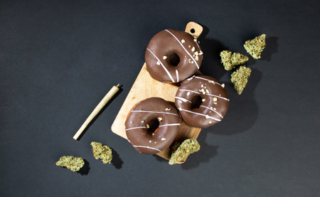 Chocolate-coated donuts with hazelnut topping lie on a wooden board among dry buds of medical marijuana, next to a joint. On a black background, flatleyの写真素材