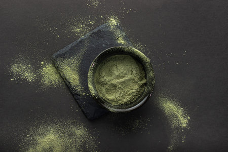 Green powder in a small ceramic bowl, on a stone stand, on a black background. The background around...