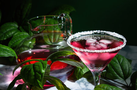 Red drink with ice in a margarita glass with a sugar rim. In the background is a jug with a cocktail, surrounded by green leaves of a plant. On a marble table. summer pastimeの写真素材
