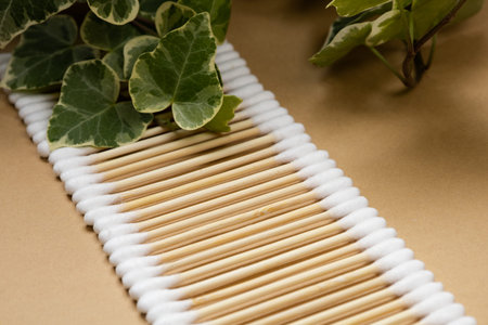 A lot of wooden cotton buds lie in a row on a beige craft paper background. Near the green leaves of...