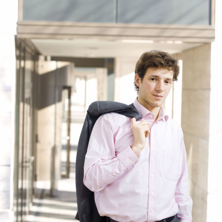 Happy young businessman standing in front of office building, effortless, shirt with open collar with jacket on shoulder, smiling.の写真素材