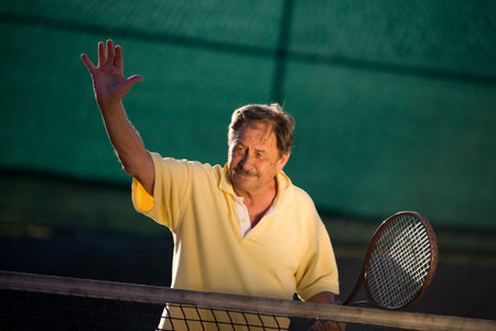 Active senior man in his 70s is posing on the tennis court with tennis racket in hand. Outdoor, sunlight.の写真素材
