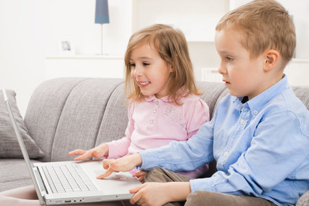 Happy children sitting on couch at home, browsing internet on laptop computer, looking at screen...
