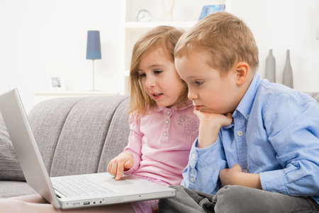 Young children sitting on couch at home, using laptop computer, looking at screen with serious...